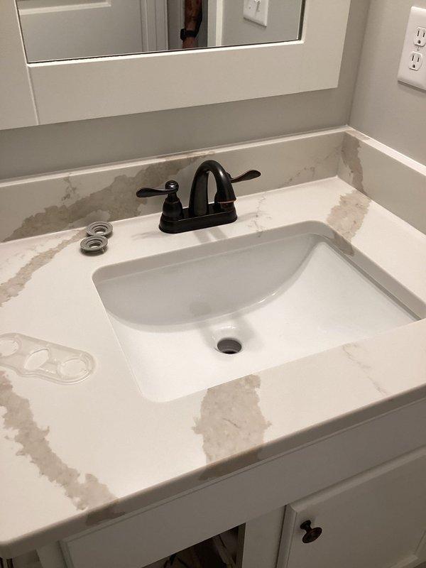Completed plumbing project estimate for bathroom vanity reinstallation following new countertop and sink installation. Assessed two vanity locations: first bathroom features dark charcoal countertop with white veining and undermount white sink with brushed nickel three-piece faucet set; second bathroom has white veined countertop with undermount rectangular sink and matte black dual-handle centerset faucet. Documented existing conditions including visible surface staining on white countertop requiring attention, confirmed all plumbing connections accessible through white cabinetry, and verified faucet mounting configurations for proper reinstallation procedures.