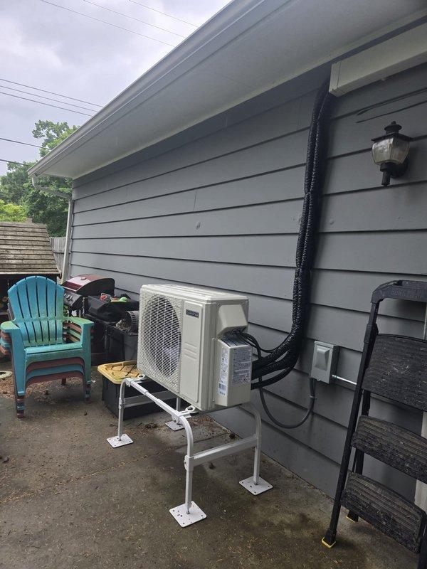 Completed installation of ductless mini-split system at residential property. Mounted outdoor condenser unit on white metal bracket stand against exterior wall with gray vinyl siding, secured refrigerant line set with black insulated cover running vertically to roofline, and installed electrical disconnect box adjacent to unit. System tested and confirmed operational upon completion.