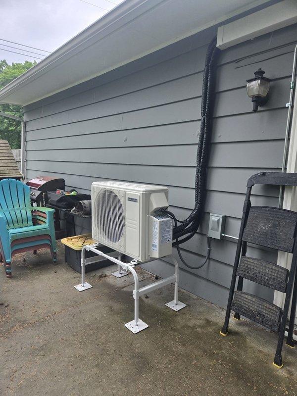 Completed installation of ductless mini-split system at residential property. Mounted outdoor condenser unit on white metal bracket stand against exterior wall with gray vinyl siding, secured refrigerant line set with black insulated cover running vertically to roofline, and installed electrical disconnect box adjacent to unit. System tested and confirmed operational upon completion.