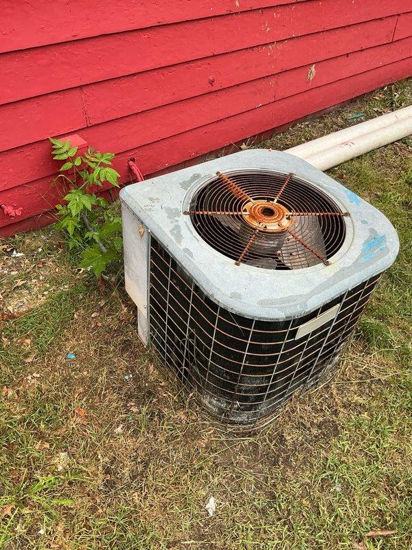 Performed comprehensive assessment of outdoor air conditioning condenser unit showing significant age-related wear, weathering, and deterioration. Unit exhibits rust spots, discoloration, and accumulated debris, with installation positioned against building exterior on uneven ground surface. Documented current equipment condition through photographic evidence and provided estimate for AC system evaluation and potential replacement or repair recommendations.