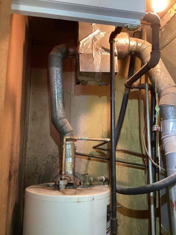 Provided estimate for replacement of 33-year-old water heater that has exceeded twice its expected lifespan and is actively leaking, causing water damage to surrounding area. Assessment included evaluation of current placement and potential relocation of unit due to space constraints and accessibility concerns. Documented existing conditions showing aged galvanized piping, deteriorated wooden framing with moisture damage, and compromised floor area requiring remediation during installation.