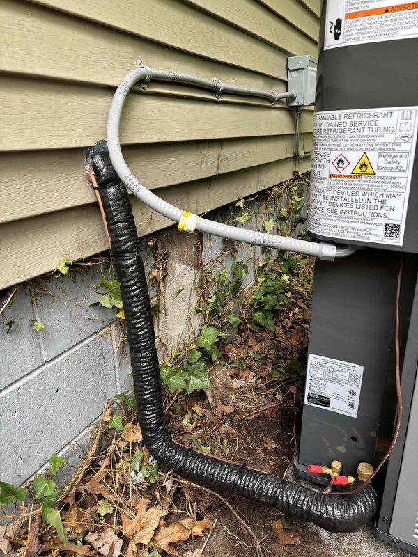 Successfully completed add-on air conditioning system installation at residential property. Installed new outdoor condensing unit with gray metal casing on concrete pad against exterior wall with beige vinyl siding. Mounted electrical disconnect box to building exterior per code requirements. Ran refrigerant line set consisting of insulated copper suction line and liquid line from condensing unit to building penetration, properly secured to siding with appropriate brackets and fasteners. Installed black flexible conduit for electrical connections. All refrigerant lines properly insulated and secured. System positioned for optimal airflow and serviceability. Installation completed per manufacturer specifications and local building codes. Unit operational and ready for service.