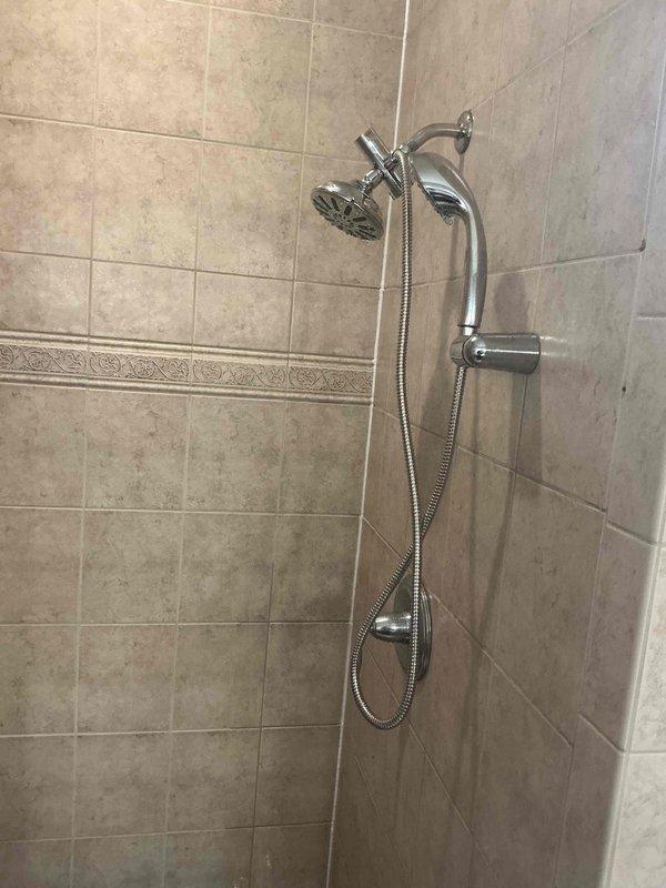Completed replacement of Kohler shower fixture components and installed new showerhead assembly in tiled shower enclosure. The installation included mounting a chrome handheld shower wand with flexible metal hose and holder bracket, along with a curved shower arm supporting a round showerhead, ensuring proper function and secure positioning on the ceramic tile wall.