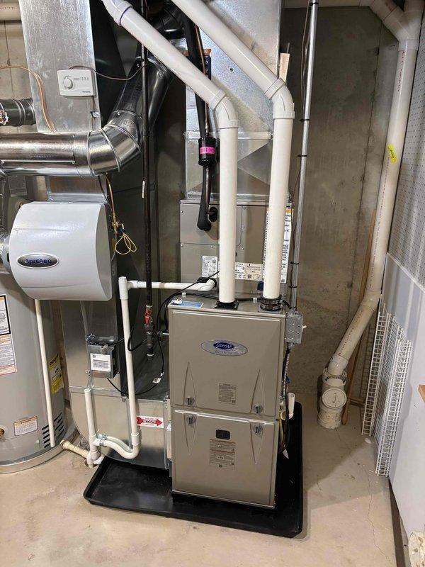 Completed full HVAC system installation including new Carrier furnace and outdoor AC condenser unit. Furnace installed in basement utility room on protective drain pan with humidifier integration, PVC venting, and proper gas line and electrical connections. Exterior condenser unit positioned on concrete pad with refrigerant lines and electrical conduit properly secured to building exterior, complete with all necessary ductwork modifications and system startup.