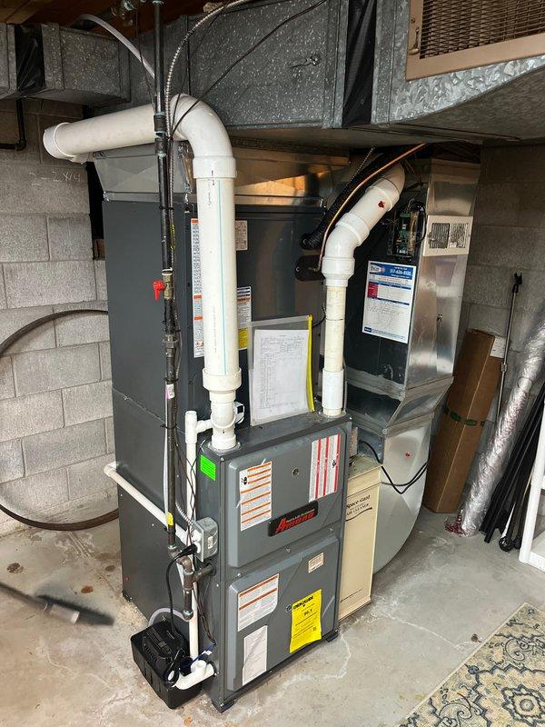 Completed full HVAC system replacement including outdoor condenser unit and indoor air handler. Installed new gray residential condenser with proper clearance from building exterior, connected electrical disconnect box with appropriate conduit routing, and positioned unit on stable pad with adequate drainage. Inside utility space, installed furnace with integrated air handler, configured white PVC venting system extending to exterior, and connected all necessary ductwork including galvanized sheet metal and flexible ducting. Verified proper electrical connections, tested refrigerant lines, confirmed condensate drainage operation, and ensured all safety labels and manufacturer specifications were clearly visible. System commissioned and tested for proper heating and cooling operation with all components functioning within normal parameters.