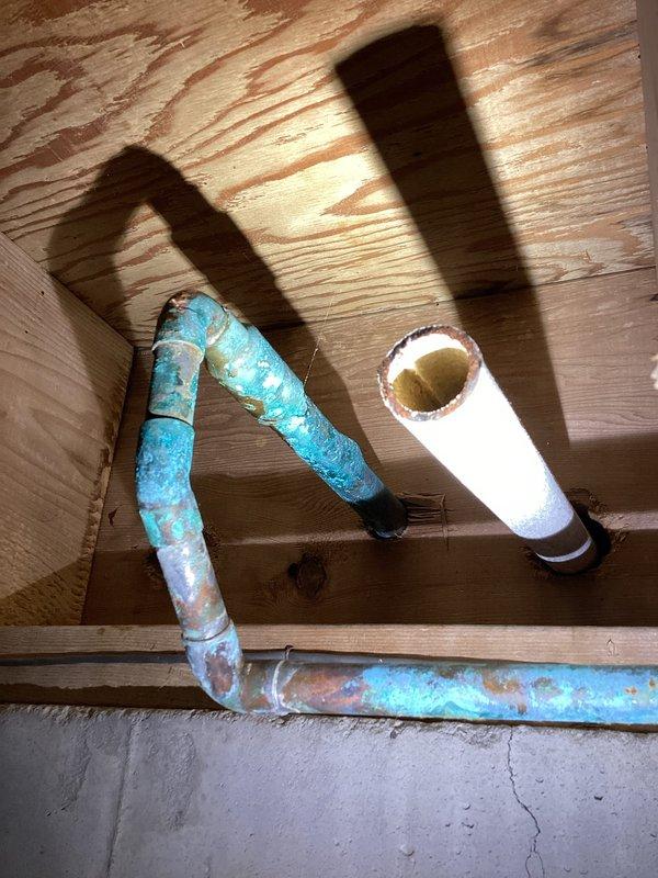 Responded to basement water intrusion occurring when exterior hose bib was in use. Upon inspection, discovered severely corroded copper supply line in basement serving outdoor spigot on 50-year-old home. The copper piping exhibited extensive turquoise and green patina throughout, indicating advanced oxidation and deterioration. Identified leak point at corroded section of horizontal pipe run beneath floor joists, which only manifested under pressure when hose was actively running. Documented condition of both the compromised basement piping and the two exterior hose bibs mounted on brick foundation wall. The affected supply line showed compromised integrity due to age-related corrosion, requiring replacement to prevent continued water intrusion into basement space during hose usage.