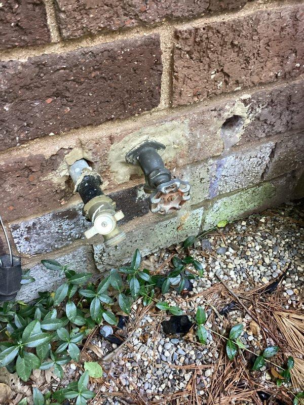 Responded to basement water intrusion occurring when exterior hose bib was in use. Upon inspection, discovered severely corroded copper supply line in basement serving outdoor spigot on 50-year-old home. The copper piping exhibited extensive turquoise and green patina throughout, indicating advanced oxidation and deterioration. Identified leak point at corroded section of horizontal pipe run beneath floor joists, which only manifested under pressure when hose was actively running. Documented condition of both the compromised basement piping and the two exterior hose bibs mounted on brick foundation wall. The affected supply line showed compromised integrity due to age-related corrosion, requiring replacement to prevent continued water intrusion into basement space during hose usage.