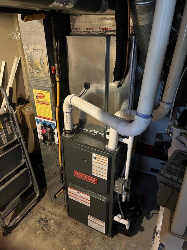 Completed full air conditioning system replacement at residential property. Removed existing AC condenser unit and installed new Goodman brand outdoor condensing unit on concrete pad in side yard location adjacent to house. Unit positioned between exterior wall with beige vinyl siding and deck structure with proper clearances maintained for airflow and service access. Connected refrigerant lines, electrical disconnect box, and white PVC condensate drain line to exterior wall. Interior installation included Goodman furnace unit in basement mechanical room with associated ductwork connections, including flexible metal ducts and insulated ductwork. Installed white PVC condensate drain and ventilation piping from furnace. Verified all electrical connections, tested system startup and operation, confirmed proper refrigerant charge and airflow. System commissioned and operational upon completion.