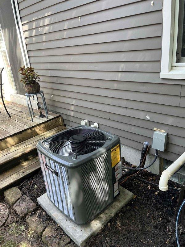 Completed full air conditioning system replacement at residential property. Removed existing AC condenser unit and installed new Goodman brand outdoor condensing unit on concrete pad in side yard location adjacent to house. Unit positioned between exterior wall with beige vinyl siding and deck structure with proper clearances maintained for airflow and service access. Connected refrigerant lines, electrical disconnect box, and white PVC condensate drain line to exterior wall. Interior installation included Goodman furnace unit in basement mechanical room with associated ductwork connections, including flexible metal ducts and insulated ductwork. Installed white PVC condensate drain and ventilation piping from furnace. Verified all electrical connections, tested system startup and operation, confirmed proper refrigerant charge and airflow. System commissioned and operational upon completion.