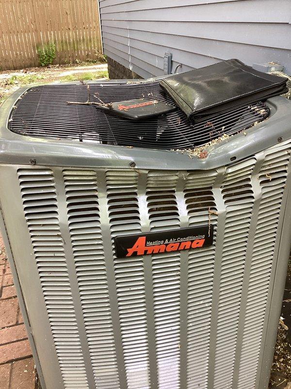 Responded to a no-cooling call where a fallen tree had struck the outdoor Amana air conditioning unit, causing significant damage to the condenser cabinet and preventing fan operation. Upon inspection, found the top grille and protective cover collapsed with visible bending of the metal housing, accumulated debris throughout the unit, and confirmed the condenser fan was unable to rotate due to structural damage. Documented the extent of damage with photographs showing the compromised cabinet, damaged top grille assembly, and internal coil components for insurance and replacement purposes.