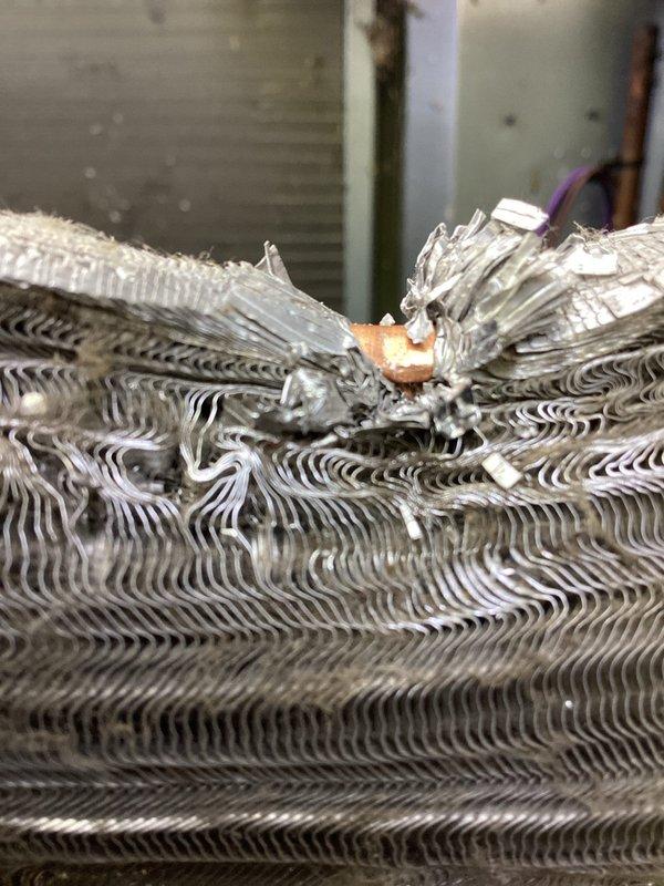 Responded to a no-cooling call where a fallen tree had struck the outdoor Amana air conditioning unit, causing significant damage to the condenser cabinet and preventing fan operation. Upon inspection, found the top grille and protective cover collapsed with visible bending of the metal housing, accumulated debris throughout the unit, and confirmed the condenser fan was unable to rotate due to structural damage. Documented the extent of damage with photographs showing the compromised cabinet, damaged top grille assembly, and internal coil components for insurance and replacement purposes.