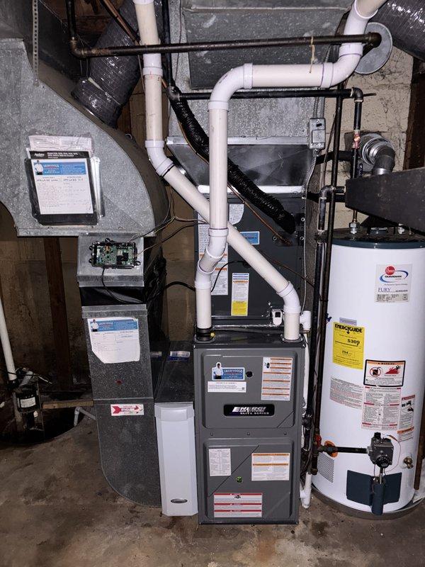 Completed full HVAC system replacement including outdoor condensing unit and indoor air handler. Removed existing equipment and installed new gray furnace unit in mechanical room with proper venting using white PVC piping throughout. Installed new outdoor AC condenser on existing concrete pad with gray metal housing and top-mounted fan. Connected black flexible ductwork to furnace and routed around piping system. Verified all electrical connections, control boards, and safety labels properly installed on equipment. Ensured proper drainage with black corrugated pipe at exterior unit location. System tested and operational upon completion.