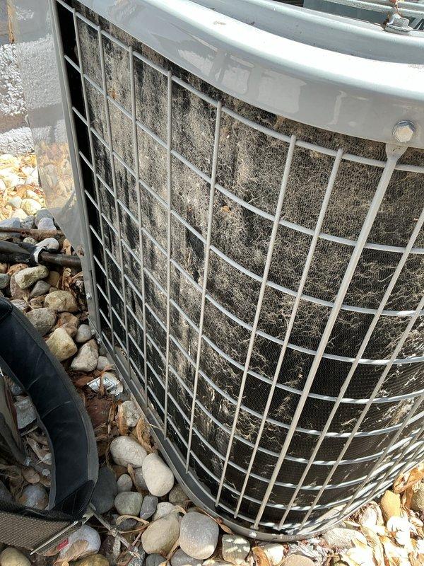 Responded to service call for air conditioning system with reported cracked condensate pan leaking water into furnace and pinched refrigerant line. Upon arrival, performed thorough inspection of outdoor condenser unit and indoor components. Found condenser unit with severely clogged protective cage, fins, and mesh filter heavily restricted with accumulated dirt, dust, and debris buildup indicating extended period without maintenance. Documented condition of drainage system at base of unit including black plastic drain pan component and concrete pad installation. Photographed extent of debris accumulation on cylindrical condenser unit which was completely obscuring internal components and significantly impacting airflow and system efficiency. Assessed reported concerns regarding condensate pan integrity and refrigerant line condition for further evaluation and repair recommendations.