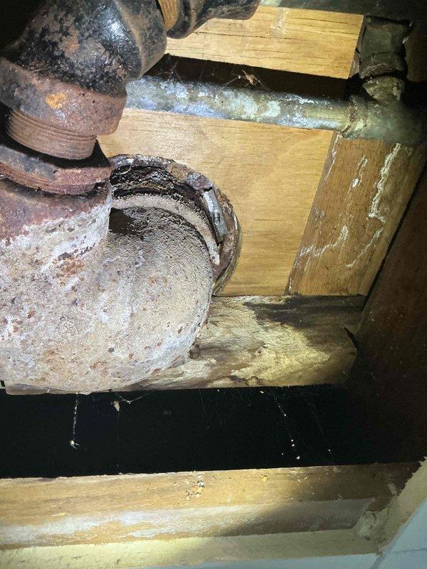 Responded to service call for toilet re-leveling and ceiling leak investigation. Upon inspection, accessed crawl space beneath bathroom and identified heavily corroded cast iron waste pipes with significant rust deterioration at threaded connections and elbow fittings. Documented existing plumbing infrastructure showing advanced corrosion, aged galvanized piping, and moisture damage to surrounding wooden floor joists. Assessed structural support members for water staining and verified integrity of pipe connections secured with metal hose clamps. Photographed deteriorated plumbing system for documentation and future reference regarding potential leak sources affecting ceiling above.