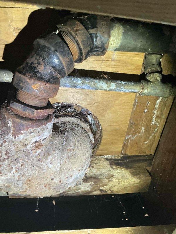 Responded to service call for toilet re-leveling and ceiling leak investigation. Upon inspection, accessed crawl space beneath bathroom and identified heavily corroded cast iron waste pipes with significant rust deterioration at threaded connections and elbow fittings. Documented existing plumbing infrastructure showing advanced corrosion, aged galvanized piping, and moisture damage to surrounding wooden floor joists. Assessed structural support members for water staining and verified integrity of pipe connections secured with metal hose clamps. Photographed deteriorated plumbing system for documentation and future reference regarding potential leak sources affecting ceiling above.