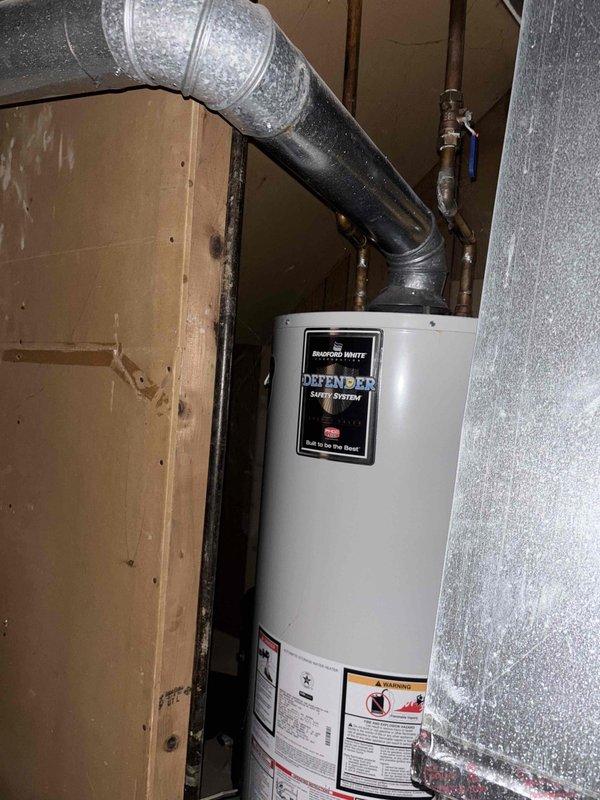Completed estimate for water heater replacement options at residential property. Evaluated existing Bradford White Defender Safety System tank-style water heater installation in basement utility area, documenting current equipment configuration including copper supply piping, blue-handled shutoff valve, and flexible vent ductwork penetrating exterior wall. Assessed installation location with exposed wall framing and provided pricing options for both traditional tank-style and tankless water heater systems to meet customer requirements.