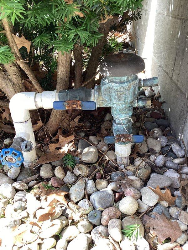 Responded to plumbing leak at outdoor irrigation system affecting a vacuum breaker valve assembly that had been leaking for approximately one week at a 39-year-old residential property. Upon arrival, inspected the backflow prevention device and associated piping located in landscaped area near building foundation. The brass vacuum breaker assembly showed significant weathering and oxidation consistent with the age of the home, with turquoise-green patina visible on copper components. Documented existing conditions with photographs showing the complete plumbing configuration including white PVC supply lines, ball valves with blue handles, and the deteriorated backflow preventer positioned among decorative river rock landscaping. Confirmed active leak at vacuum breaker valve connection points and assessed overall condition of outdoor water management system for repair recommendations.