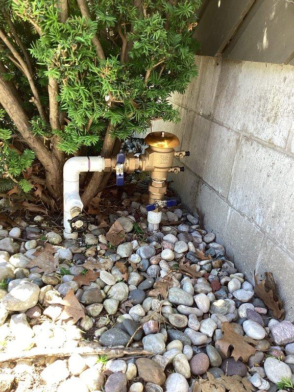 Responded to plumbing leak at outdoor irrigation system affecting a vacuum breaker valve assembly that had been leaking for approximately one week at a 39-year-old residential property. Upon arrival, inspected the backflow prevention device and associated piping located in landscaped area near building foundation. The brass vacuum breaker assembly showed significant weathering and oxidation consistent with the age of the home, with turquoise-green patina visible on copper components. Documented existing conditions with photographs showing the complete plumbing configuration including white PVC supply lines, ball valves with blue handles, and the deteriorated backflow preventer positioned among decorative river rock landscaping. Confirmed active leak at vacuum breaker valve connection points and assessed overall condition of outdoor water management system for repair recommendations.