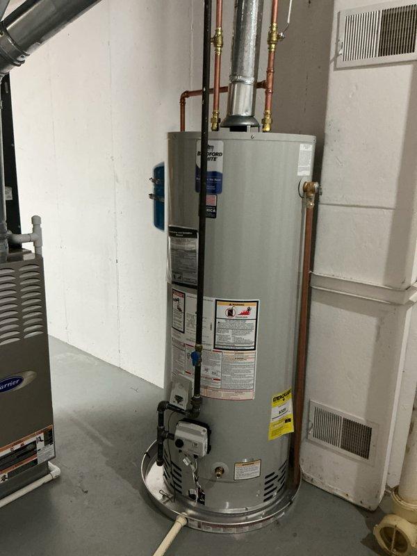 Completed installation of a 50-gallon natural gas water heater in basement utility room. Unit was positioned against concrete block wall with proper clearances maintained for ventilation and service access. Connected copper supply lines with brass fittings to existing plumbing infrastructure and installed blue expansion tank above water heater to accommodate thermal expansion in closed system. Ran gas line connection to unit and installed PVC temperature and pressure relief valve discharge pipe to floor drain per code requirements. Verified all connections for leaks, tested gas valve operation, and confirmed proper venting. System was filled, purged of air, and brought up to operating temperature with all safety controls functioning correctly.