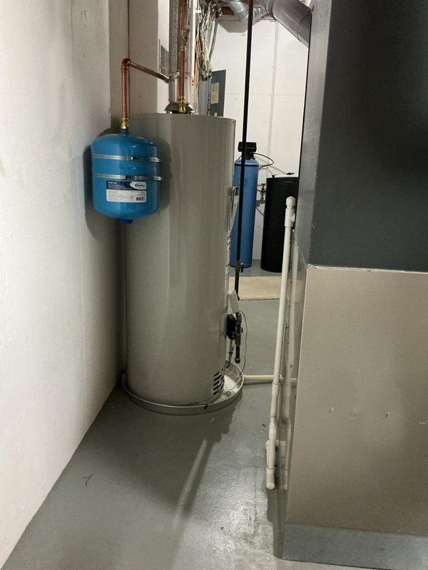 Completed installation of a 50-gallon natural gas water heater in basement utility room. Unit was positioned against concrete block wall with proper clearances maintained for ventilation and service access. Connected copper supply lines with brass fittings to existing plumbing infrastructure and installed blue expansion tank above water heater to accommodate thermal expansion in closed system. Ran gas line connection to unit and installed PVC temperature and pressure relief valve discharge pipe to floor drain per code requirements. Verified all connections for leaks, tested gas valve operation, and confirmed proper venting. System was filled, purged of air, and brought up to operating temperature with all safety controls functioning correctly.