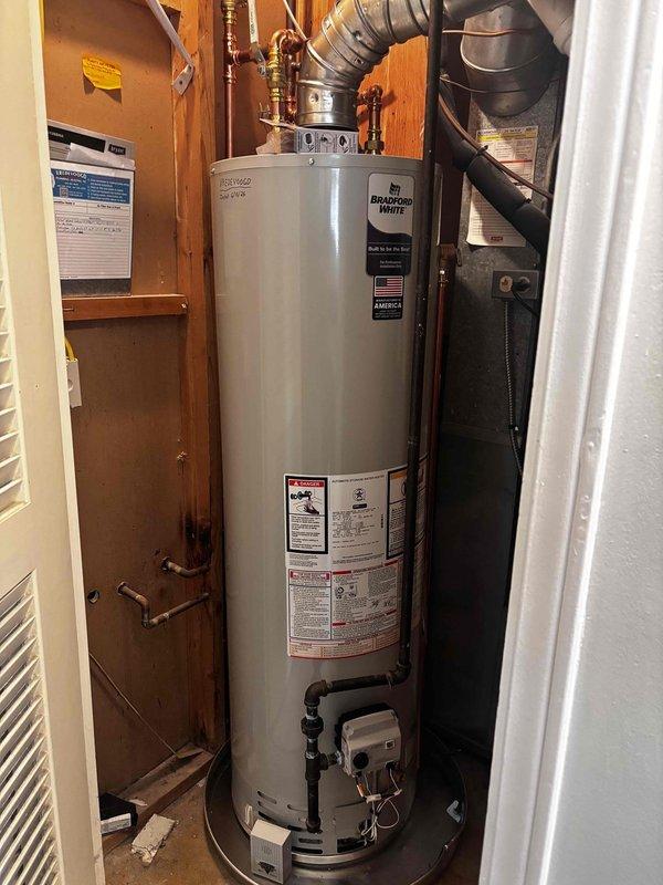 Completed installation of Bradford White natural draft 40-gallon water heater in residential basement utility area. Removed existing unit and installed new slim-profile model with proper copper supply line connections, brass shut-off valves, and metallic vent ductwork. Connected black PVC drain piping to code-compliant relief valve assembly, mounted unit on elevated platform, and verified all plumbing connections, gas lines, and ventilation system for proper operation and safety compliance.