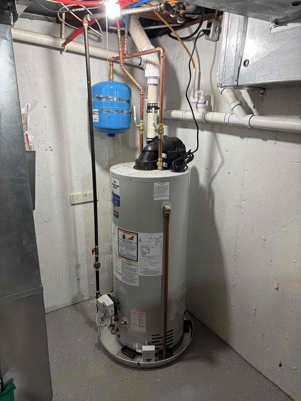 Completed full water heater installation in residential utility room. Installed new cylindrical water heater with all necessary plumbing connections, including copper piping with proper hot and cold water lines, brass fittings, and shut-off valves. Mounted blue expansion tank to wall with appropriate copper pipe connections to system. Installed black circulation pump on top of unit and connected all required PVC drainage piping. Verified all connections for proper operation and tested system functionality upon completion.