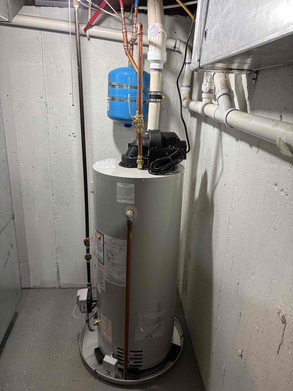 Completed full water heater installation in residential utility room. Installed new cylindrical water heater with all necessary plumbing connections, including copper piping with proper hot and cold water lines, brass fittings, and shut-off valves. Mounted blue expansion tank to wall with appropriate copper pipe connections to system. Installed black circulation pump on top of unit and connected all required PVC drainage piping. Verified all connections for proper operation and tested system functionality upon completion.