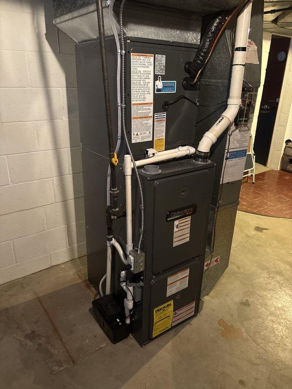 Completed full HVAC system replacement including indoor air handler and outdoor condensing unit. Installed new furnace and air handler assembly in basement utility area against existing cinder block wall, ensuring proper mounting and structural support. Routed white PVC condensate drain lines and venting pipes along left side of unit with professional organization. Connected black flexible conduit for electrical service between upper and lower components. Verified all manufacturer labels and safety warnings are visible and intact on equipment front panels. Established proper ductwork connections to existing metal supply plenum at ceiling level.

Positioned new outdoor condensing unit on level concrete pad adjacent to home foundation. Installed unit on stable mounting surface with adequate clearance from vinyl siding and basement window for proper airflow and future service access. Completed refrigerant line connections between indoor and outdoor components. Tested system startup, verif