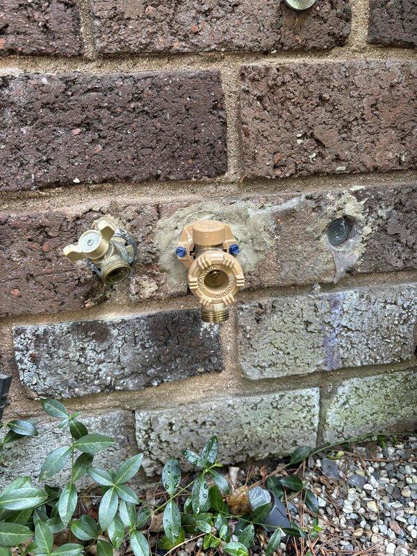 Responded to follow-up service call for persistent leak at exterior hose bib installation. Upon arrival, inspected three brass hose bibs mounted on exterior brick wall and associated plumbing components beneath concrete foundation. Located source of ongoing leak at pressure regulator valve assembly with blue adjustment screws. Tightened connections at regulator body and verified proper seating of valve components. Tested all three hose bib fixtures under pressure to ensure complete water-tight seal. Confirmed leak resolution and proper operation of all exterior water connections.
