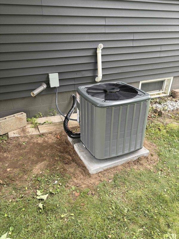 Completed full HVAC system replacement including outdoor condensing unit and indoor air handler with evaporator coil. Installed new condenser on existing concrete pad adjacent to foundation with proper clearances maintained. Ran refrigerant linesets through exterior wall penetration with copper lines insulated appropriately. Mounted electrical disconnect box on exterior siding and installed black flexible conduit for power supply to outdoor unit. Installed white PVC condensate drain line routed vertically along exterior wall. Indoor air handler positioned above existing furnace in basement utility area between wall studs. Connected refrigerant lines with brown insulation jacket running vertically alongside equipment. Installed black flexible supply ductwork from air handler secured with metal clamps. Routed white PVC drain lines and completed electrical connections at indoor unit including circuit breaker installation. System tested for proper operation, refrigerant charge verified, an