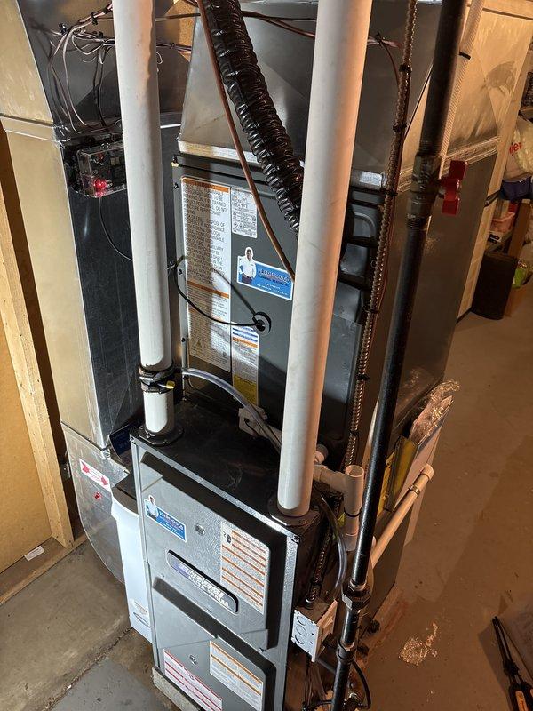 Completed full HVAC system replacement including outdoor condensing unit and indoor air handler with evaporator coil. Installed new condenser on existing concrete pad adjacent to foundation with proper clearances maintained. Ran refrigerant linesets through exterior wall penetration with copper lines insulated appropriately. Mounted electrical disconnect box on exterior siding and installed black flexible conduit for power supply to outdoor unit. Installed white PVC condensate drain line routed vertically along exterior wall. Indoor air handler positioned above existing furnace in basement utility area between wall studs. Connected refrigerant lines with brown insulation jacket running vertically alongside equipment. Installed black flexible supply ductwork from air handler secured with metal clamps. Routed white PVC drain lines and completed electrical connections at indoor unit including circuit breaker installation. System tested for proper operation, refrigerant charge verified, an