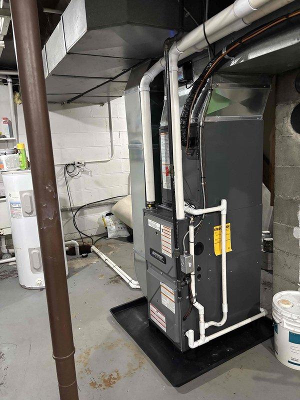 Completed full installation of residential furnace and air conditioning system in basement mechanical room. New HVAC system includes modern air handler unit with proper PVC condensate drainage lines, ventilation piping, and black drain pan installation on concrete floor. All equipment properly connected to existing ductwork, electrical service, and plumbing connections with appropriate safety labels and manufacturer specifications verified. System tested and operational upon completion.