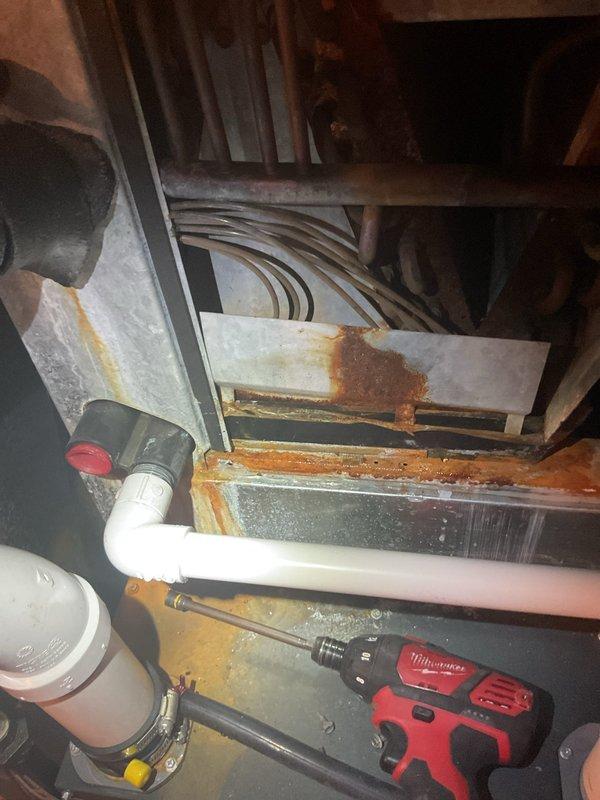 Responded to service call for reported refrigerant line leak at approximately 20-year-old air conditioning system. Upon inspection, accessed basement mechanical space and located the condensate drain line and refrigerant piping leading to outdoor AC unit. Extensive corrosion and rust damage was observed on metal ductwork and surrounding components, indicating prolonged moisture exposure in the area. Documented existing conditions with photographs showing deteriorated metal surfaces, PVC drain piping, copper refrigerant lines, and overall system configuration. The reported leak appears to be related to compromised piping connections or condensate drainage issues based on visible water damage patterns and mineral deposits on ductwork surfaces. Further diagnostic work and repair recommendations were provided to address the moisture intrusion and prevent additional system damage.
