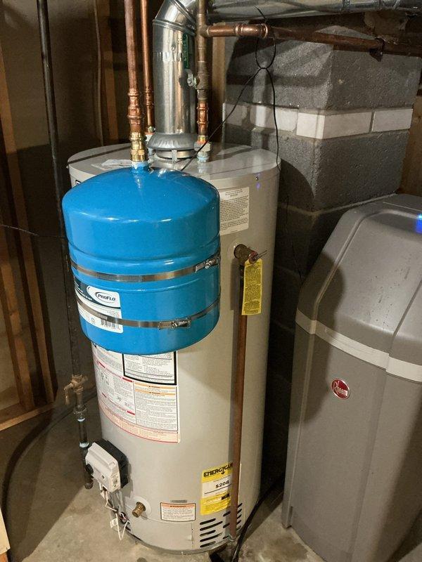 Installed domestic hot water expansion tank on existing water heater system in basement utility area. Mounted blue expansion tank to top of water heater using proper metal clamps and connected to system via copper piping with brass fittings to protect against thermal expansion and maintain system pressure compliance.