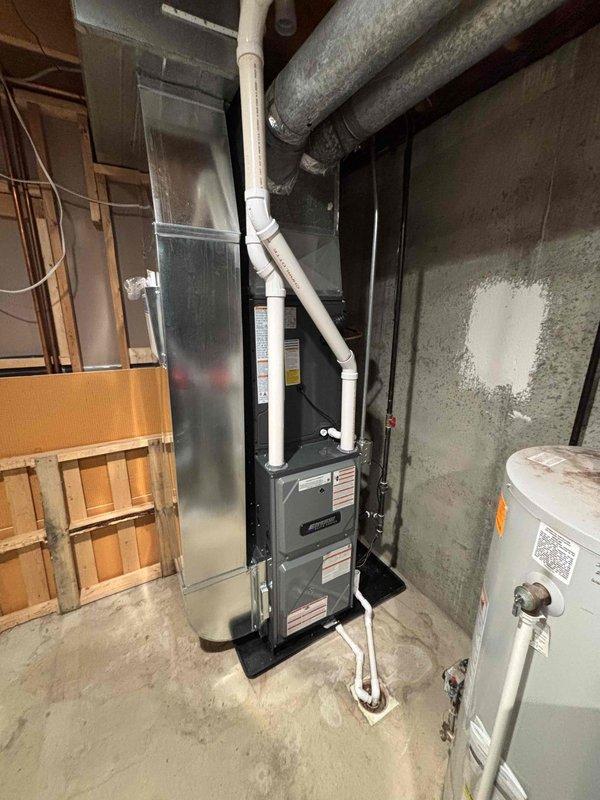 Completed full installation of new furnace and air conditioning system in basement utility area. Installed gray metal furnace unit with proper venting using white PVC piping routed upward and connected to insulated ductwork system. Configured refrigerant lines with appropriate valving, secured black piping to concrete wall with mounting brackets, and positioned equipment on protective base pad within mechanical closet. All components installed per manufacturer specifications with proper clearances, connections verified, and system tested for operational readiness.