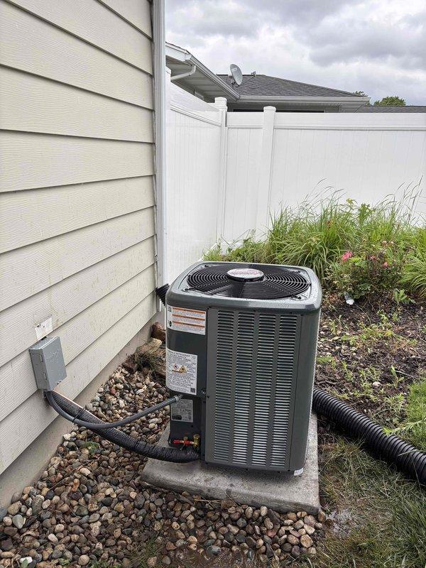 Complete air conditioning system replacement performed at residential property. Removed existing outdoor condenser unit and installed new AC condenser on concrete pad against exterior wall with proper clearances. Unit positioned on decorative rock base with adequate drainage provisions. Connected electrical disconnect box to house siding per code requirements. Installed black corrugated condensate drain line with proper routing away from equipment. Verified all manufacturer labels and safety stickers properly affixed to unit panels. Tested system operation including condenser fan function and confirmed proper startup. Installation completed with unit secured on level concrete pad, all electrical connections made to code, and surrounding landscaping preserved.