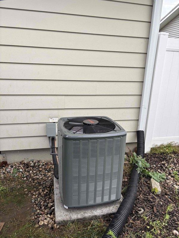 Complete air conditioning system replacement performed at residential property. Removed existing outdoor condenser unit and installed new AC condenser on concrete pad against exterior wall with proper clearances. Unit positioned on decorative rock base with adequate drainage provisions. Connected electrical disconnect box to house siding per code requirements. Installed black corrugated condensate drain line with proper routing away from equipment. Verified all manufacturer labels and safety stickers properly affixed to unit panels. Tested system operation including condenser fan function and confirmed proper startup. Installation completed with unit secured on level concrete pad, all electrical connections made to code, and surrounding landscaping preserved.