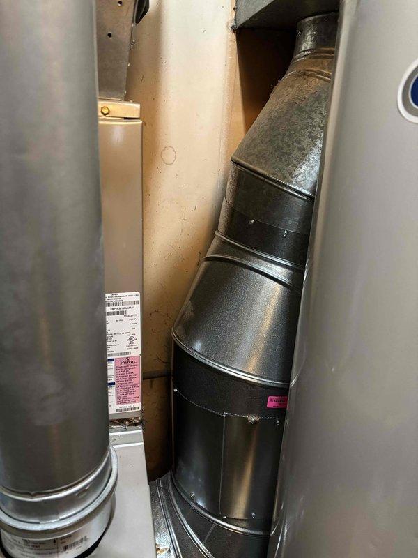 Completed full furnace installation in residential utility closet. Installed new Carrier-brand furnace unit with proper clearances for maintenance access. Connected vertical flue piping and integrated ductwork to existing HVAC system. Secured all mounting brackets and verified proper ventilation. Installation includes complete connection of supply and return air systems with black metal ductwork routing. All manufacturer specifications followed for equipment placement adjacent to existing water heater. System installed on concrete pad with appropriate spacing per code requirements.