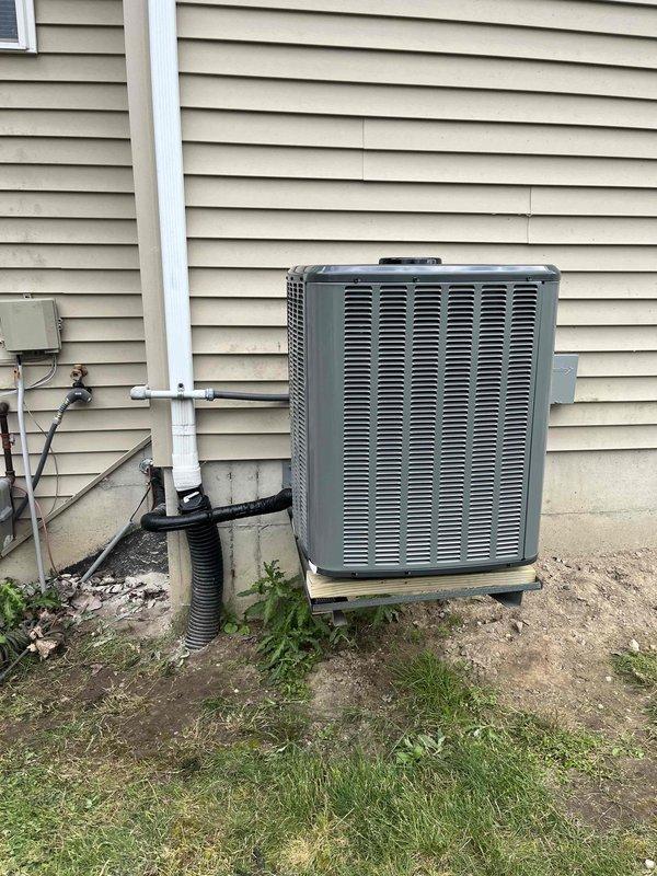 Complete residential air conditioning system replacement performed. Removed existing outdoor condenser unit and indoor evaporator coil assembly. Installed new outdoor condensing unit on existing concrete pad against exterior wall with beige vinyl siding, ensuring proper clearances for airflow and service access. Unit positioned with adequate ventilation through horizontal louvers and secured to pad per manufacturer specifications.

Inside utility area, installed new evaporator coil assembly atop existing furnace unit. Ran new refrigerant line set consisting of black insulated copper lines from outdoor condenser to indoor coil, routing lines along foundation and through wall penetration. Secured all refrigerant lines with appropriate mounting brackets and hangers. Completed brazing connections at both indoor and outdoor units, pressure tested system, and evacuated to manufacturer specifications.

Connected electrical supply to outdoor unit using appropriate gauge wiring and conduit. Ver