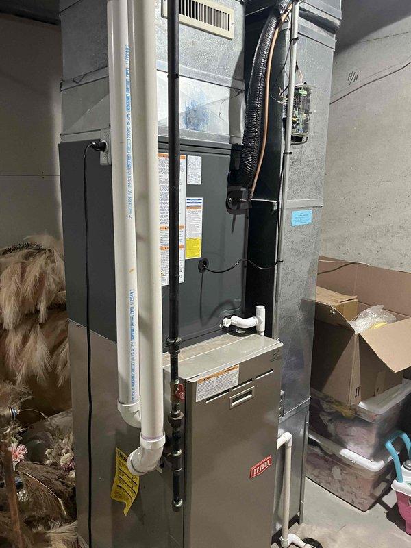 Complete residential air conditioning system replacement performed. Removed existing outdoor condenser unit and indoor evaporator coil assembly. Installed new outdoor condensing unit on existing concrete pad against exterior wall with beige vinyl siding, ensuring proper clearances for airflow and service access. Unit positioned with adequate ventilation through horizontal louvers and secured to pad per manufacturer specifications.

Inside utility area, installed new evaporator coil assembly atop existing furnace unit. Ran new refrigerant line set consisting of black insulated copper lines from outdoor condenser to indoor coil, routing lines along foundation and through wall penetration. Secured all refrigerant lines with appropriate mounting brackets and hangers. Completed brazing connections at both indoor and outdoor units, pressure tested system, and evacuated to manufacturer specifications.

Connected electrical supply to outdoor unit using appropriate gauge wiring and conduit. Ver