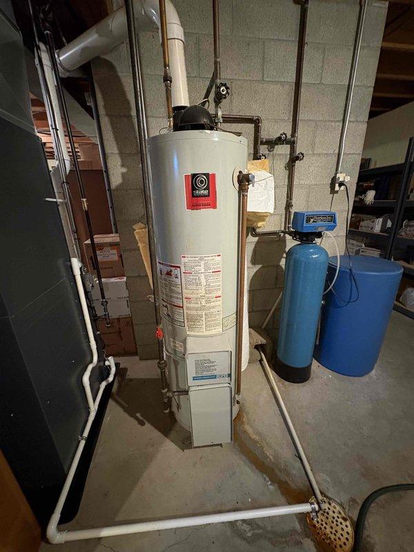 Provided water heater replacement estimate for 25-year-old unit located in basement utility area. Inspected existing white cylindrical water heater on raised concrete platform with copper and PVC connections, including white PVC exhaust vent and gas lines. Documented current installation configuration including proximity to blue water softener system and furnace unit, assessed routing of supply and exhaust piping through ceiling space to upper levels, and photographed all relevant mechanical connections for replacement planning.