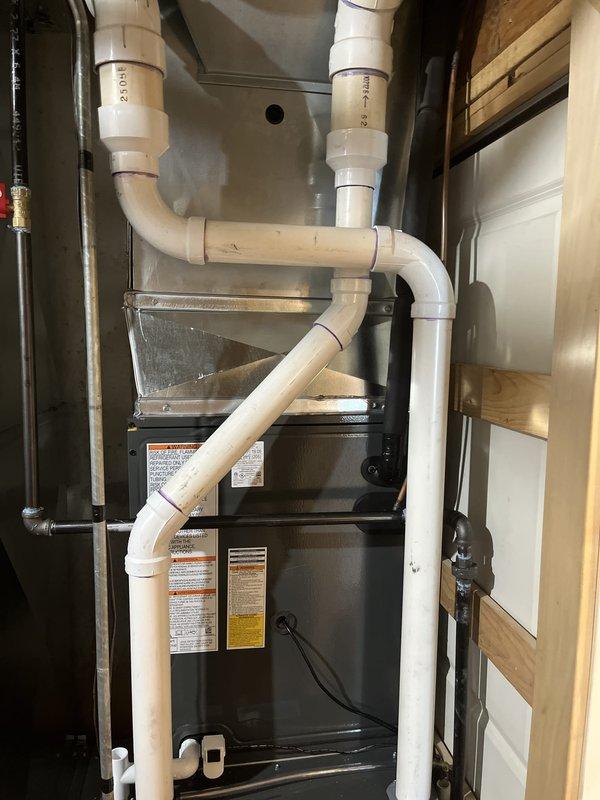 Completed full residential HVAC system installation including furnace and air conditioning unit. Installed dark-colored vertical furnace in utility closet with complete PVC venting and drainage network, including multiple vertical and angled pipe configurations for proper exhaust and condensate management. Mounted outdoor condensing unit on concrete pad with proper electrical disconnect, conduit routing, and refrigerant line connections to house exterior. All manufacturer labels, safety compliance stickers, and energy guide documentation properly affixed to equipment. System installed per manufacturer specifications with secure mounting, proper clearances, and complete electrical connections verified.