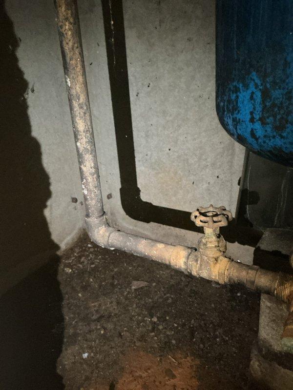 Responded to emergency plumbing leak at residential property with well pump system. Upon arrival, inspected basement utility area and identified leak originating from piping connected to well pump assembly. Located corroded manual valve with wheel handle on horizontal pipe run near floor level, showing significant age-related deterioration and mineral buildup. Assessed condition of blue pressure tank exhibiting surface corrosion and paint failure. Examined sump pump basin with rusted cover and PVC standpipe, noting moisture accumulation and sediment in basin area. Documented existing conditions through photographs of plumbing infrastructure, valve assembly, pressure tank, and sump system. Addressed immediate leak concern at well pump connection point to prevent further water damage to basement area and restore proper system operation.