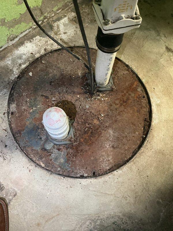 Responded to emergency plumbing leak at residential property with well pump system. Upon arrival, inspected basement utility area and identified leak originating from piping connected to well pump assembly. Located corroded manual valve with wheel handle on horizontal pipe run near floor level, showing significant age-related deterioration and mineral buildup. Assessed condition of blue pressure tank exhibiting surface corrosion and paint failure. Examined sump pump basin with rusted cover and PVC standpipe, noting moisture accumulation and sediment in basin area. Documented existing conditions through photographs of plumbing infrastructure, valve assembly, pressure tank, and sump system. Addressed immediate leak concern at well pump connection point to prevent further water damage to basement area and restore proper system operation.