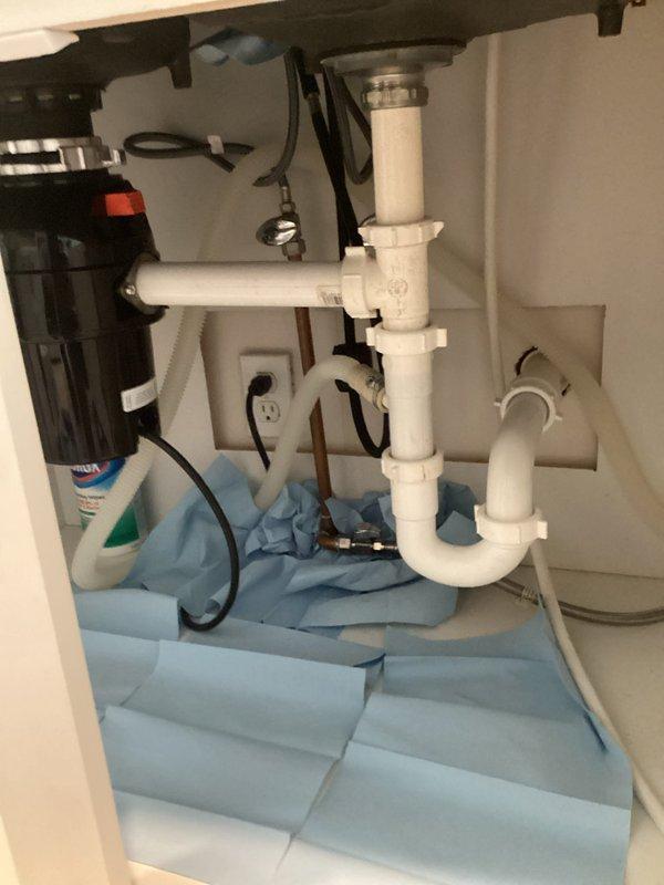 Responded to kitchen cabinet leak beneath double-basin sink. Upon inspection, identified moisture issue in cabinet base and installed absorbent pads to protect cabinet floor while addressing plumbing concerns. Inspected PVC P-trap assembly, drain connections, and garbage disposal unit for potential leak sources. Verified all pipe connections were secure and properly sealed. Photographed plumbing configuration and cabinet conditions for documentation purposes. System tested and monitored to ensure proper drainage function and confirm leak resolution.