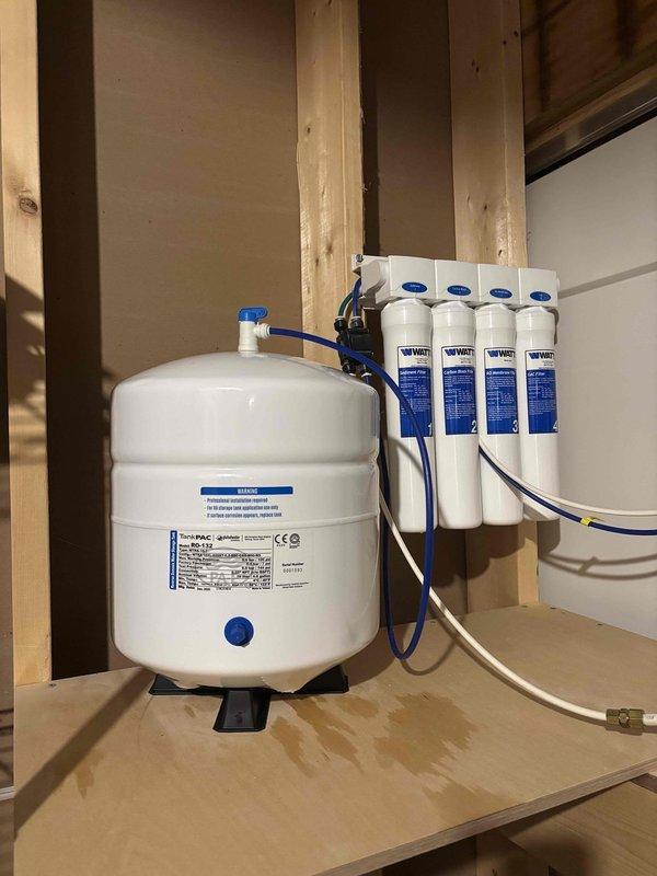 Performed diagnostic evaluation of residential water softener system and documented existing plumbing configuration. Inspected under-sink plumbing connections including garbage disposal unit and PVC drainage system to assess potential water supply issues. Examined water filtration system with pressurized storage tank and four-stage filter cartridge assembly mounted in utility area, verifying connections and component condition. Documented system layout with photographs for scheduled follow-up repair work requiring 1-2 hours to complete necessary adjustments and restore proper water softener operation.