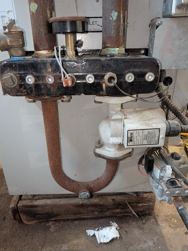 Completed comprehensive boiler tune-up on aging heating system with visible corrosion on manifold assembly and copper piping connections. Performed standard maintenance procedures including inspection of black rectangular manifold unit, circulator pump motor, U-bend copper piping, and all electrical connections; system operational upon completion despite advanced age and surface oxidation on components.