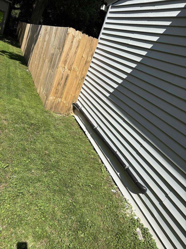 Completed installation of add-on air conditioning condenser unit at residential property. Unit was positioned on concrete pad against exterior wall with proper clearances maintained, electrical conduit and refrigerant lines connected and secured to structure, and condensate drainage system installed with flexible drain line routed away from foundation.