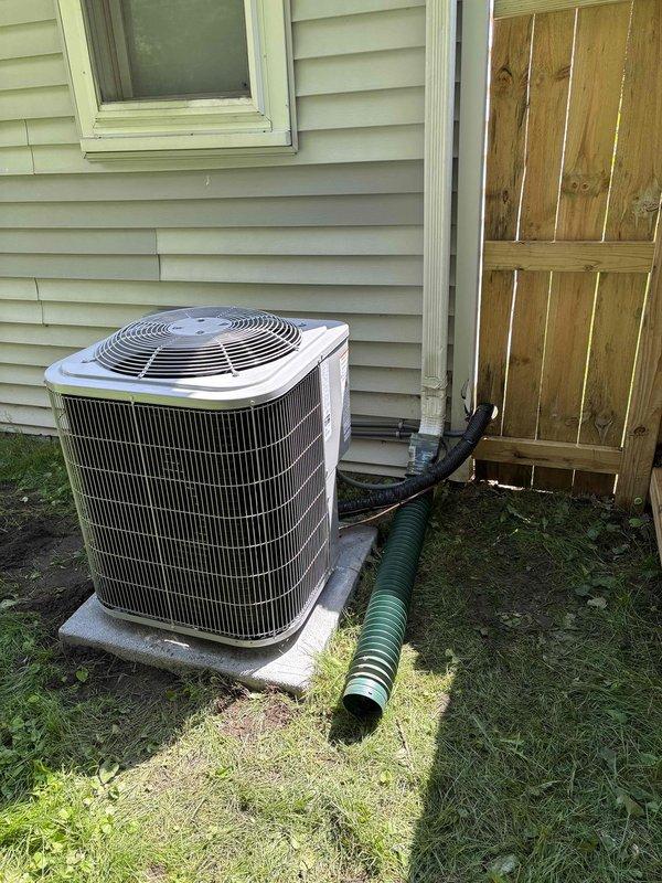 Completed installation of add-on air conditioning condenser unit at residential property. Unit was positioned on concrete pad against exterior wall with proper clearances maintained, electrical conduit and refrigerant lines connected and secured to structure, and condensate drainage system installed with flexible drain line routed away from foundation.