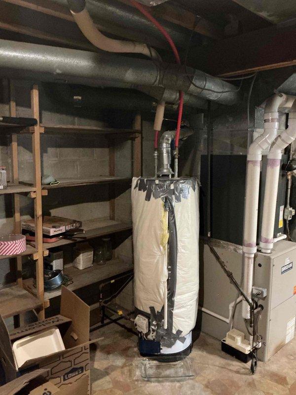 Responded to residential plumbing leak in basement utility area involving water heater discharge. Upon arrival, identified active dripping from temperature and pressure relief (TPR) valve discharge pipe onto concrete floor. Inspected water heater installation, noting unit wrapped in reflective insulation blanket with red-handled shutoff valves and various copper and PVC connections at top of tank. Examined TPR valve operation and discharge piping configuration to determine source of leak. Documented equipment condition and leak location with photographs showing water heater positioned in drain pan, adjacent furnace system, and overhead ductwork. Assessed floor area for water damage and verified proper drainage. Completed leak diagnosis and provided recommendations for necessary repairs to eliminate water discharge issue.